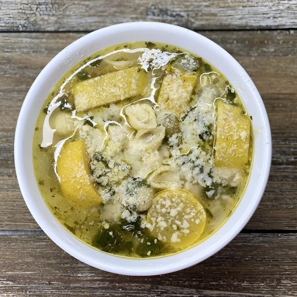 A close-up of a hearty soup with pasta, vegetables, and grated cheese in a white bowl.
