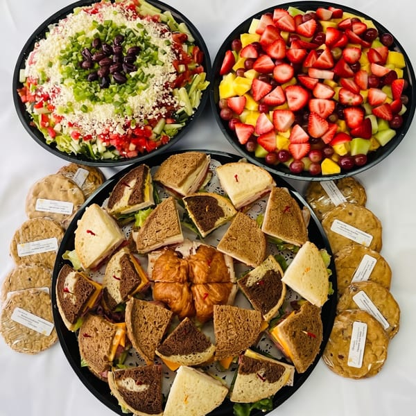 A catering spread with a large salad, fruit platter, sandwich tray, and individually wrapped cookies.
