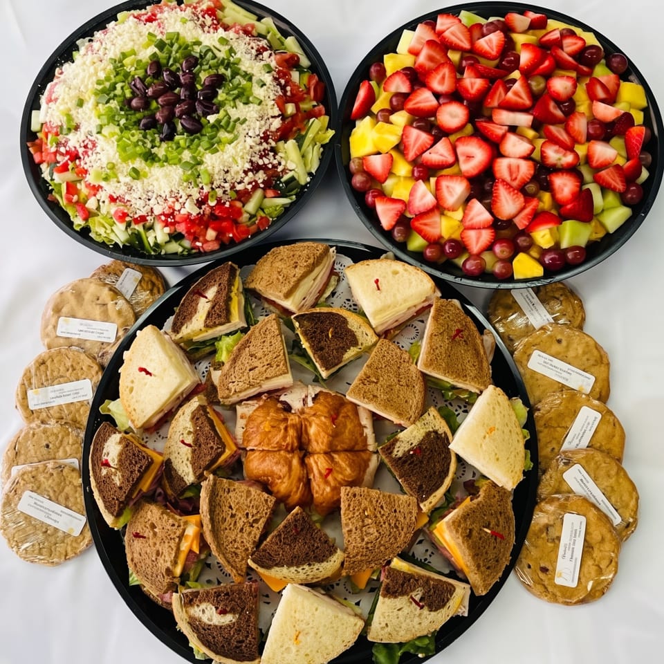 A catering spread with a large salad, fruit platter, sandwich tray, and individually wrapped cookies.