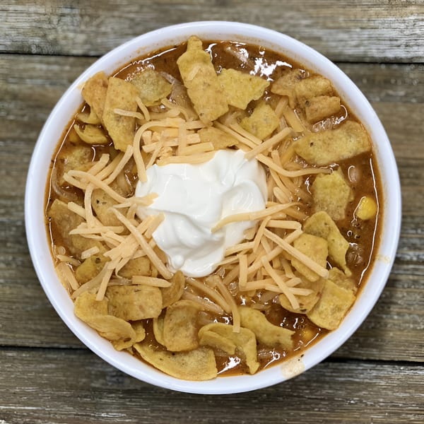 A bowl of Woody's Famous Salads chili with shredded cheese, sour cream, and corn chips.