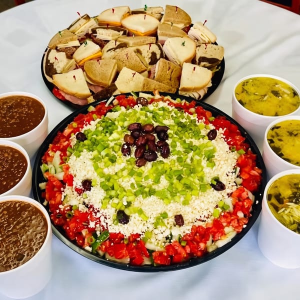 A catering spread with a platter of sandwiches, a large layered dip, and cups of soup.
