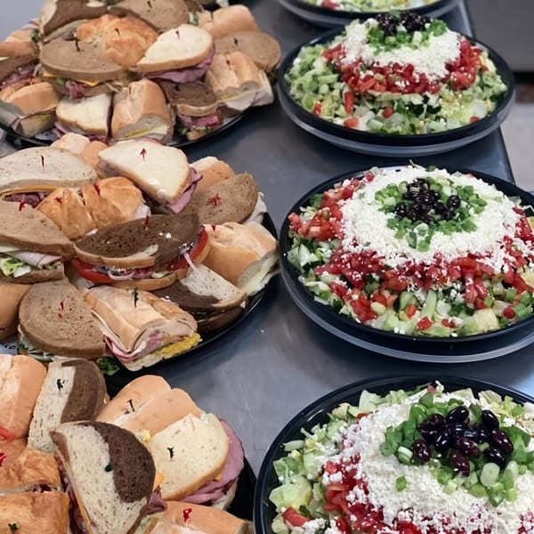 Trays of assorted sandwiches and large salads with lettuce, tomatoes, feta, and olives.